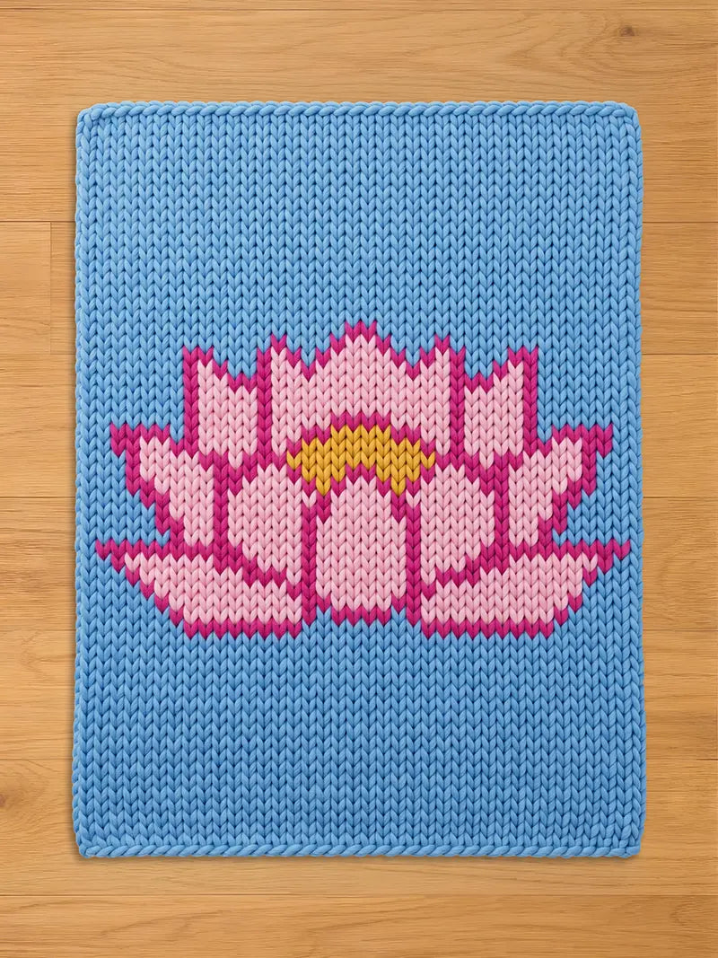 Rectangular chunky knit blanket with a light blue background, featuring a large, central lotus flower design in light pink with dark pink outlines and a yellow core, laid flat on a wooden floor.