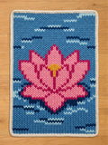 A rectangular chunky knit blanket. The design features a vibrant, multi-tonal pink lotus flower with a yellow center, set against a light blue background with dark blue and white stitched water ripples. The blanket is finished with a thick white knit border