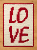 A knitted chunky blanket with the word "LOVE" in red letters on an off-white background. The letters are large and blocky, with a red border. The item is likely a decorative pillow or blanket.
