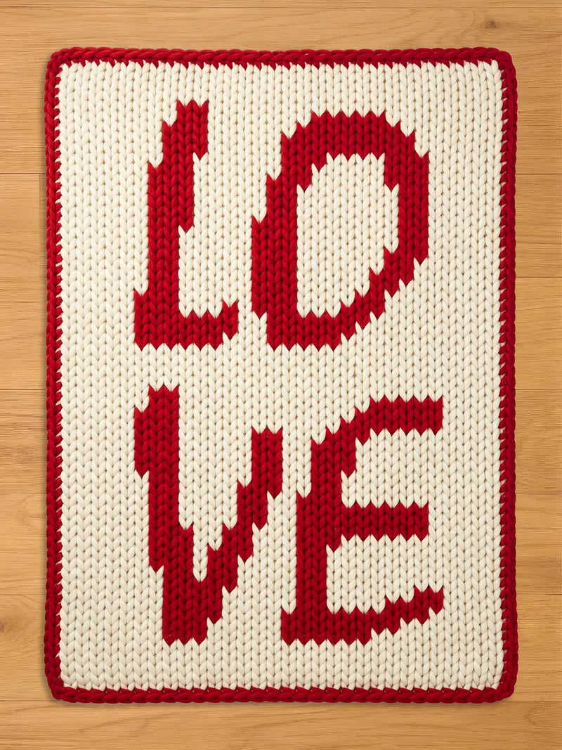 A knitted chunky blanket with the word "LOVE" in red letters on an off-white background. The letters are large and blocky, with a red border. The item is likely a decorative pillow or blanket.