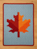 Cozy chunky knit blanket with a fall-themed red and orange maple leaf pattern, light blue background, and red knit edging.
