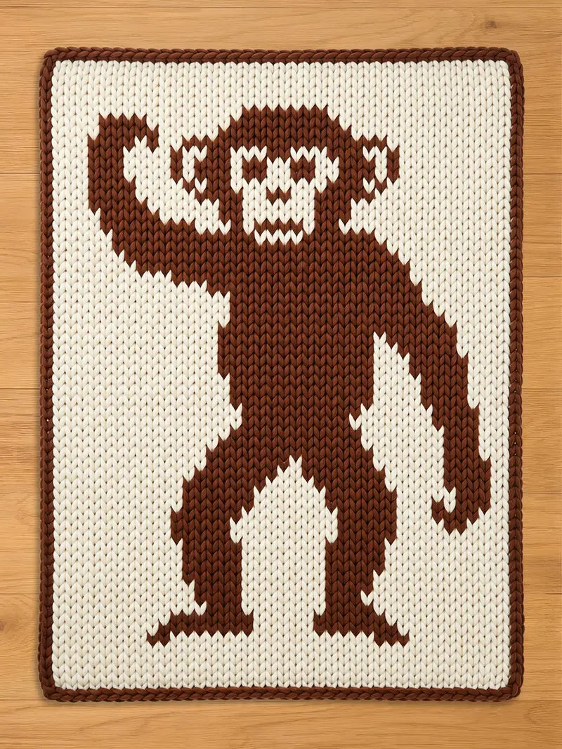 Rectangular chunky knit blanket in cream and dark brown. The center features a full-body, pixel-art style monkey with one arm raised, knitted in brown yarn. The blanket has a matching dark brown border and is displayed flat on a light wood floor