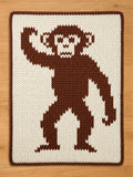 Rectangular chunky knit blanket in cream and dark brown. The center features a full-body, pixel-art style monkey with one arm raised, knitted in brown yarn. The blanket has a matching dark brown border and is displayed flat on a light wood floor