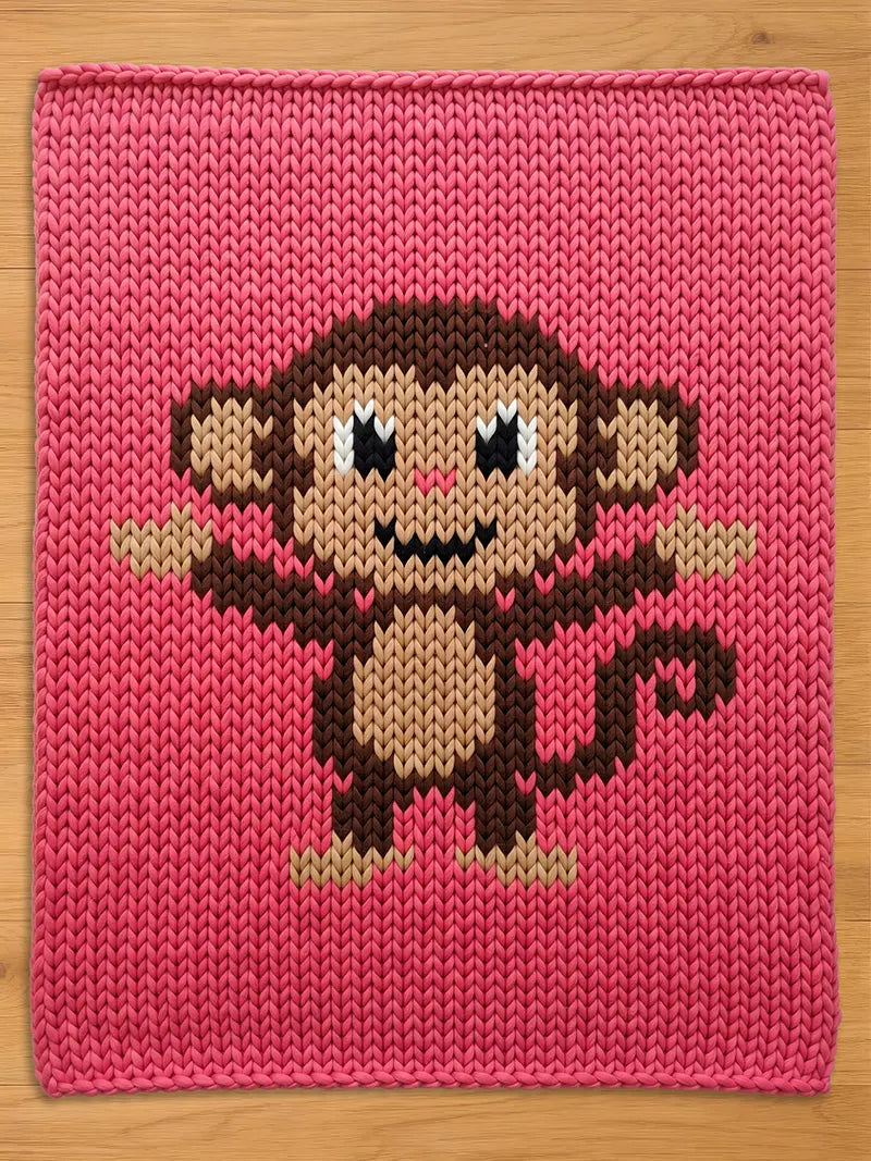 A pink knitted chunky blanket featuring a pixelated monkey design. The monkey has brown and tan fur, black eyes, and a small smile. The background is a solid, textured pink.