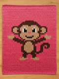 A pink knitted chunky blanket featuring a pixelated monkey design. The monkey has brown and tan fur, black eyes, and a small smile. The background is a solid, textured pink.