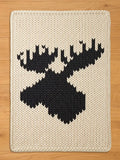 Cream-colored knitted throw blanket featuring a large black moose silhouette. The blanket has a thick, textured appearance with a detailed, geometric design of the moose head and antlers. The edges are neatly finished with a knitted border.
