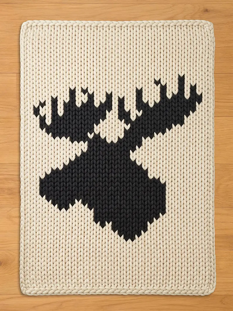 Cream-colored knitted throw blanket featuring a large black moose silhouette. The blanket has a thick, textured appearance with a detailed, geometric design of the moose head and antlers. The edges are neatly finished with a knitted border.