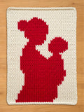 A chunky knit cream blanket featuring a large red silhouette of a mother holding a child, laid flat on a light wooden floor