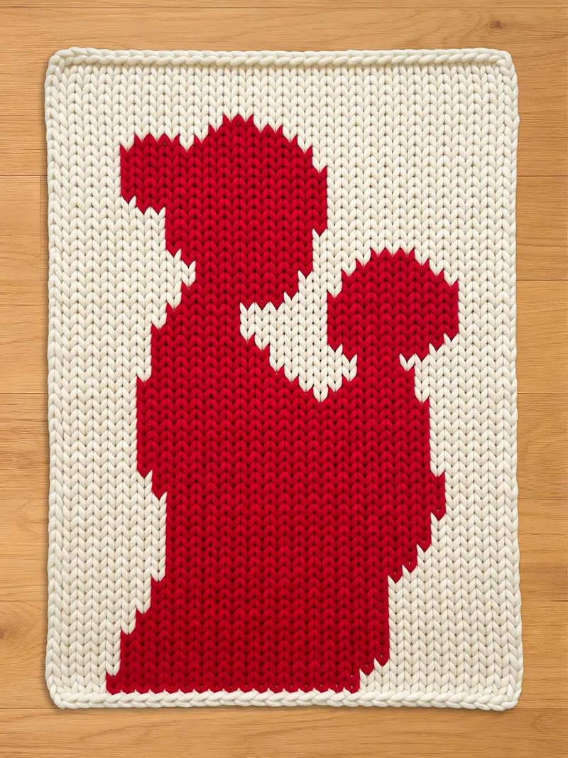 A chunky knit cream blanket featuring a large red silhouette of a mother holding a child, laid flat on a light wooden floor