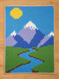 A chunky knit blanket featuring a colorful outdoor landscape design with a bright yellow sun, purple and blue snow-capped mountains, green fields, and a winding blue river