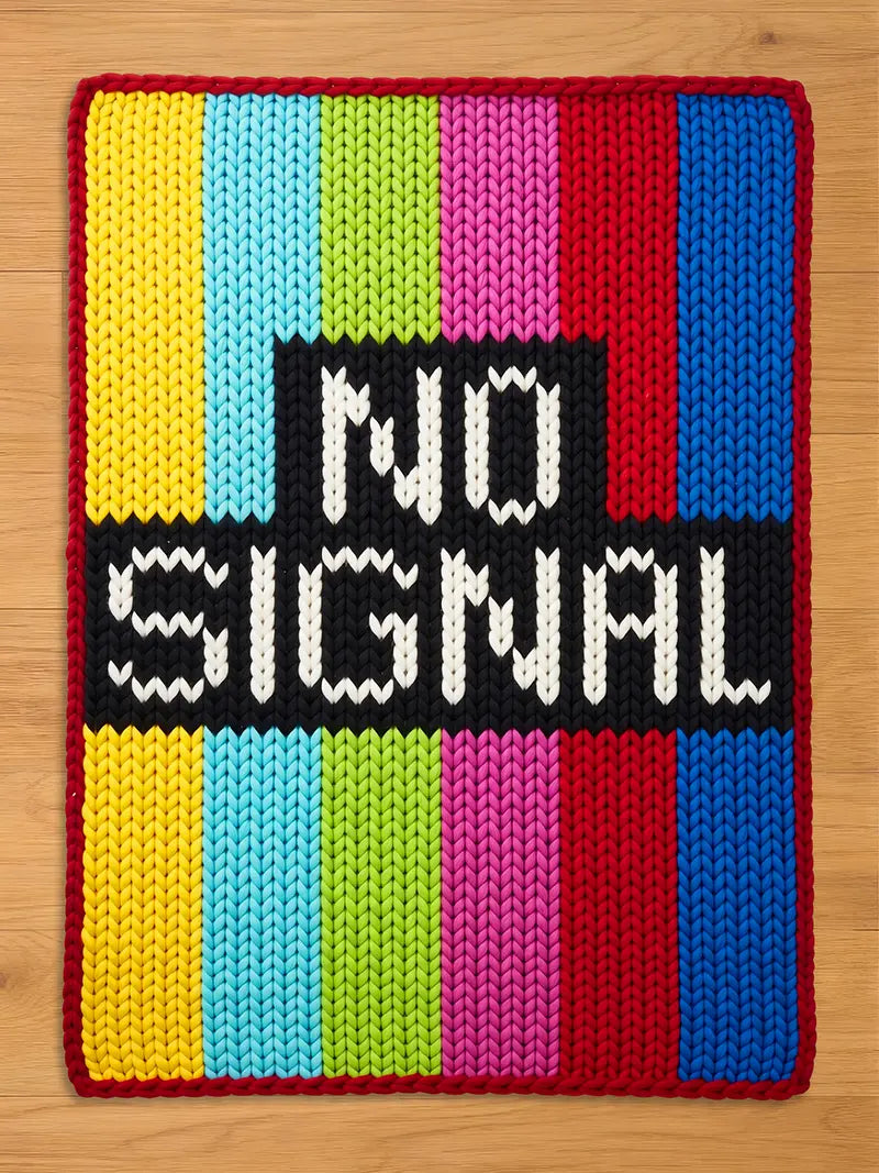 A knitted chunky blanket with a colorful striped pattern in yellow, light blue, green, pink, red, and dark blue. The rug has a black rectangle in the center with the words "NO SIGNAL" in white.
