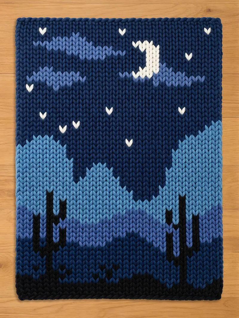 A rectangular knit blanket featuring a nighttime desert landscape. The design displays dark blue mountains, two black saguaro cacti, and a night sky with a white crescent moon and small white stars against a deep blue background.