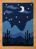 A rectangular knit blanket featuring a nighttime desert landscape. The design displays dark blue mountains, two black saguaro cacti, and a night sky with a white crescent moon and small white stars against a deep blue background.