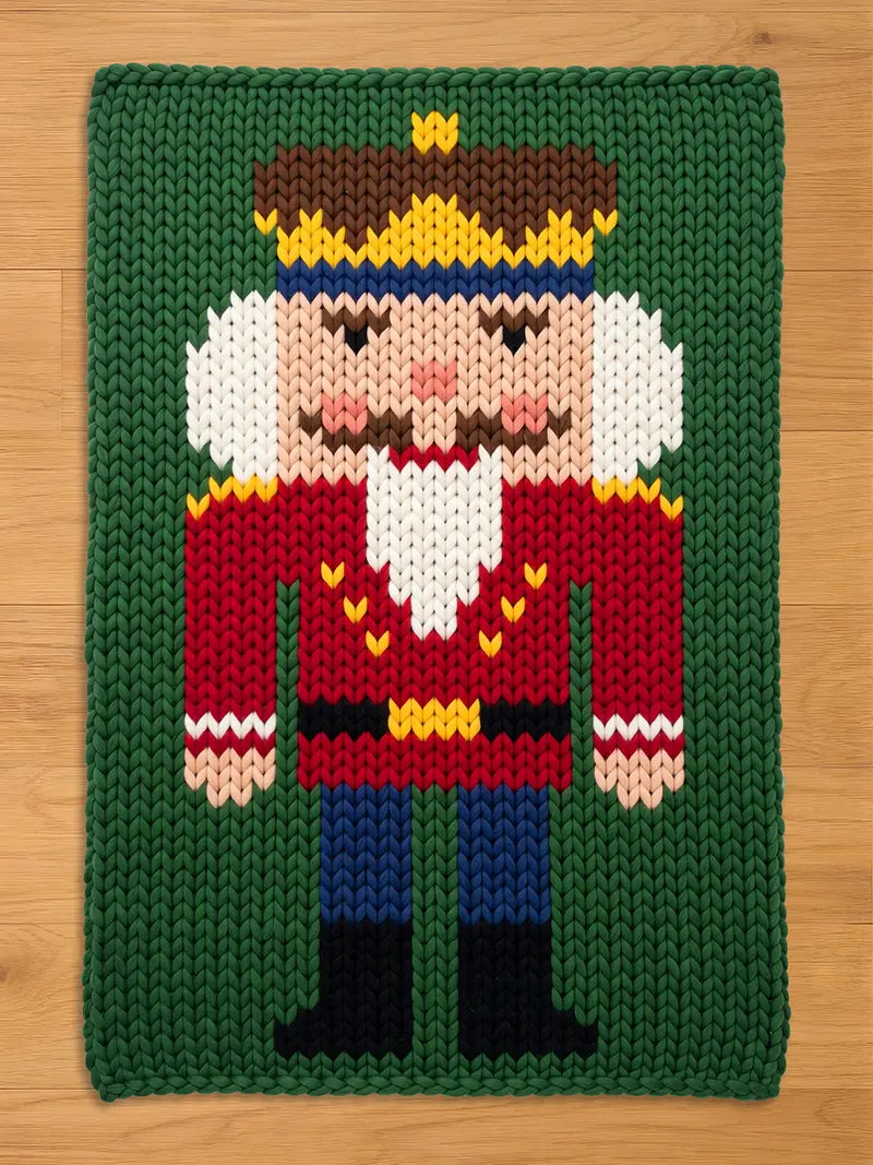 A knitted Christmas-themed rug featuring a nutcracker design. The nutcracker has a red jacket, blue pants, and a brown and yellow crown. The background is green, and the rug is rectangular.