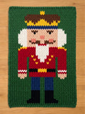 A knitted Christmas-themed rug featuring a nutcracker design. The nutcracker has a red jacket, blue pants, and a brown and yellow crown. The background is green, and the rug is rectangular.
