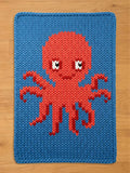 A knitted blue blanket featuring a red octopus design. The octopus has a round head, eight tentacles, and white and black eyes. The blanket is made of a thick, textured yarn.