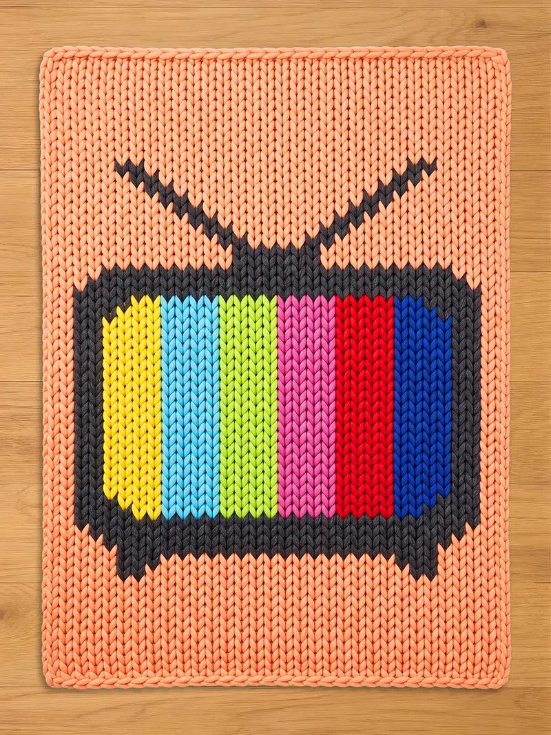 A square knitted blanket featuring a retro television screen design with the classic color test bars (yellow, light blue, green, pink, red, and blue) on a peach-orange background, laid flat on a light wood floor.