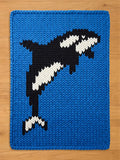 A knitted blue blanket featuring a black and white orca whale design. The orca is depicted with its distinctive black and white markings, set against a solid blue background. The blanket has a textured, woven appearance.