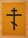 A rectangular, knitted chunky blanket in a mustard yellow color. A black, Eastern Orthodox cross is centered on the rug. The blanket has a thick, textured appearance, with visible knit stitches.