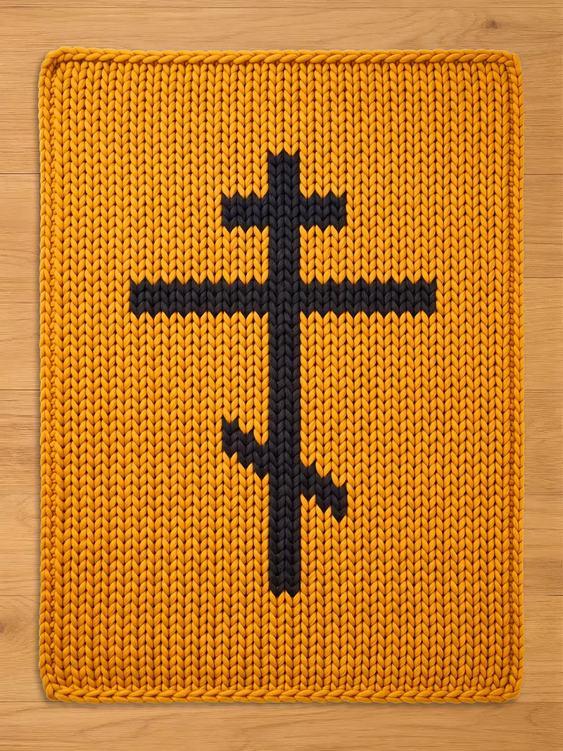 A rectangular, knitted chunky blanket in a mustard yellow color. A black, Eastern Orthodox cross is centered on the rug. The blanket has a thick, textured appearance, with visible knit stitches.