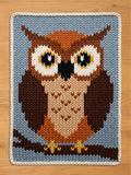 A chunky knitted blanket featuring a stylized owl design, laid flat on a light wood surface. The knit pattern shows a blue-gray background with a brown owl, cream and orange facial details, and yellow accents, finished with a cream border.