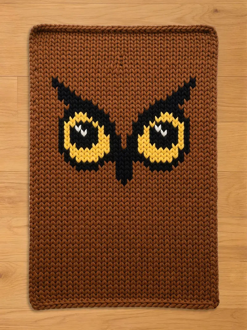 Brown knitted owl-themed chunky blanket. The blanket features a detailed owl face with yellow and black eyes, a black beak, and a brown background. The blanket is rectangular with a textured, knitted appearance.