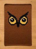 Brown knitted owl-themed chunky blanket. The blanket features a detailed owl face with yellow and black eyes, a black beak, and a brown background. The blanket is rectangular with a textured, knitted appearance.