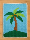 A rectangular hand-knitted chunky blanket featuring a tropical palm tree design on a light blue background, with a cream-colored braided border, laid flat on a light wood floor.