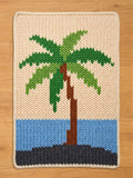 A rectangular hand-knitted chunky blanket featuring a tropical palm tree design on a sandy beige background, with blue ocean and a dark gray shore, laid flat on a light wood floor.