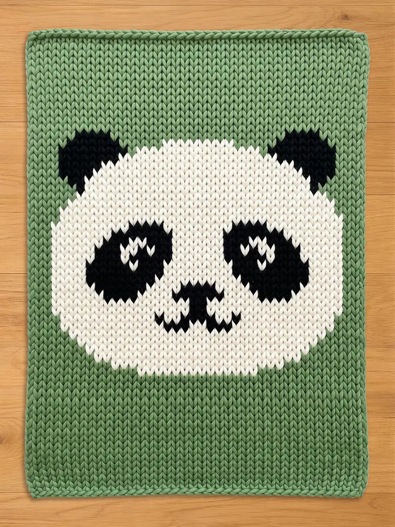 A knitted chunky blanket featuring a panda bear design. The panda's face is white with black eyes, ears, and markings, set against a green background. The item is made of a knitted material.