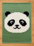 A knitted chunky blanket featuring a panda bear design. The panda's face is white with black eyes, ears, and markings, set against a green background. The item is made of a knitted material.