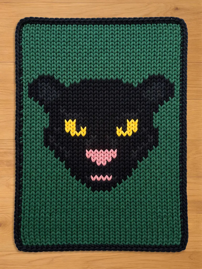 A knitted chunky blanket featuring a stylized black panther face with yellow eyes and a pink nose on a dark green background, laid flat on a light wood surface.