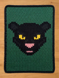 A knitted chunky blanket featuring a stylized black panther face with yellow eyes and a pink nose on a dark green background, laid flat on a light wood surface.