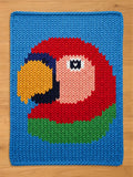 A knitted chunky blanekt featuring a colorful parrot design. The parrot has a red head, a yellow beak, and a green and blue body, set against a blue background. The piece is made with a visible knit stitch.