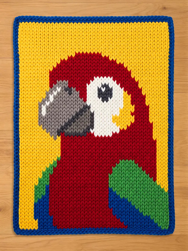 A hand-knitted rectangular chunky blanket featuring a colorful parrot design on a yellow background with a blue border, laid flat on a light wood floor.