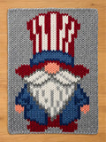 A rectangular chunky knit blanket in light gray, featuring a large patriotic gnome design in the center. The knitted gnome has a tall red and white striped hat with a dark blue brim, a round peach nose, a long white beard with gray textured lines, a dark blue coat, small peach hands, and red boots.