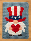 A rectangular chunky knit blanket featuring a gnome graphic. The gnome wears a tall red, white, and blue striped hat with white stars on a blue band. The gnome has a white beard, a peach-colored nose, and holds a large red heart.