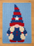 A rectangular knit blanket featuring a light blue background with a central gnome graphic. The gnome wears a tall, pointed hat with horizontal red and blue stripes and a white star, a red brim, beige beard, and blue overalls.