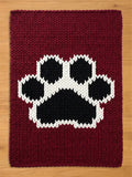 A burgundy knitted chunky blanket featuring a large paw print design in black and white. The paw print is centered on the rectangular rug, with a thick, textured knit pattern throughout.