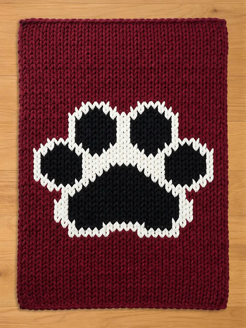 A burgundy knitted chunky blanket featuring a large paw print design in black and white. The paw print is centered on the rectangular rug, with a thick, textured knit pattern throughout.