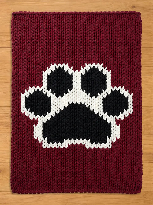 A burgundy knitted chunky blanket featuring a large paw print design in black and white. The paw print is centered on the rectangular rug, with a thick, textured knit pattern throughout.