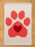 A rectangular, off-white knitted chunky blanket featuring a large paw print design in shades of red. The paw print includes a heart shape in the center. The blanket has a textured appearance and a simple, clean border.