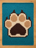 A knitted  chunky blanket with a teal background and a large paw print design. The paw print is made up of brown, tan, and cream-colored yarn, creating a textured and cozy appearance. The blanket is likely for a pet.