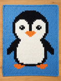 A knitted chunky blanket featuring a large penguin design. The penguin has a white body, black head and wings, orange feet and beak, and is set against a light blue background. The blanket is made of thick yarn.