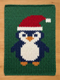 A knitted green chunky blanket featuring a penguin wearing a Santa hat. The penguin has a white belly, blue body, and orange feet. The Santa hat is red with a white pom-pom. The blanket is made of a thick, textured yarn.