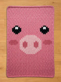 A pink knitted chunky blanket featuring a pig face design. The pig's face includes black and white eyes, a light pink snout with two nostrils, and pink cheeks. The blanket is made of a soft, textured material.