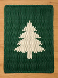 Dark green chunky knit blanket featuring a large, off-white pine tree silhouette pattern in the center, laid flat on a wooden surface