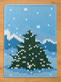 A rectangular knit blanket featuring a large dark green pine tree centered against a light blue sky with white snow-capped mountains in the background and small white stitches scattered throughout to represent falling snowflakes.