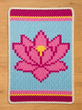 A knitted rectangular blanket featuring a pink lotus flower on a light blue background. The blanket has a cream-colored border and bands of pink, yellow, and magenta at the top and bottom. The lotus flower has a yellow center.