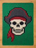 A rectangular knitted blanket featuring a central graphic of a skull wearing a pirate hat and a red bandana. The design uses cream, black, dark gray, and red yarn against a vibrant green background, showcasing a chunky knit texture.